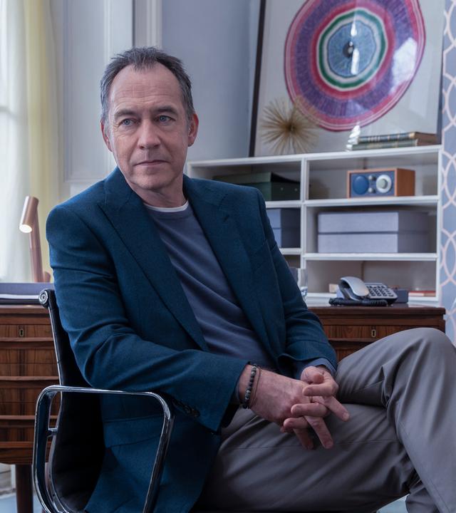 Iain Frankland (Dominic Mafham) sitzt in seinem Büro vor einem Fenster. Er trägt ein Jackett und blickt leicht zur Seite.