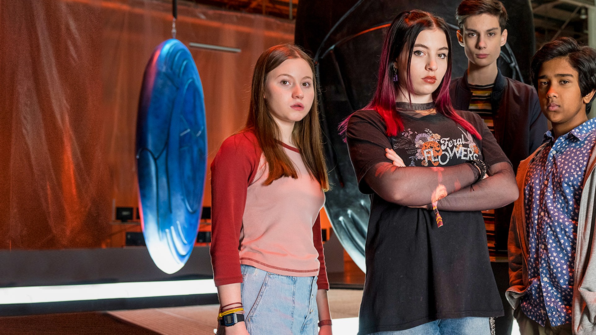 Von links nach rechts: Meg, Kaz, Louis und Glen stehen in einer Lagerhalle. Hinter ihnen sind drei sehr große sogenannte Artefakte zu sehen. Sie leuchten in verschiedenen Farben und haben verschiedene geometrische Muster auf der Oberfläche. Alle schauen entschlossen in die Kamera. Kaz hat die Arme vor der Brust verschränkt.