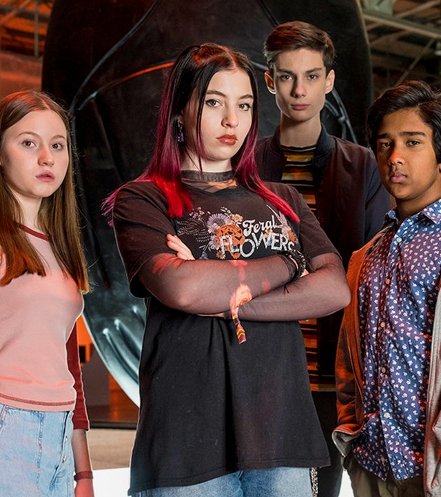 Von links nach rechts: Meg, Kaz, Louis und Glen stehen in einer Lagerhalle. Hinter ihnen sind drei sehr große sogenannte Artefakte zu sehen. Sie leuchten in verschiedenen Farben und haben verschiedene geometrische Muster auf der Oberfläche. Alle schauen entschlossen in die Kamera. Kaz hat die Arme vor der Brust verschränkt.