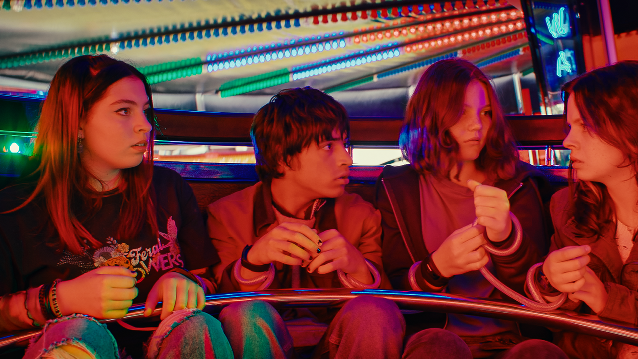 Auf einem Jahrmarkt in einem Wagen eines Karussells sitzen Kaz, Jay, Isabel und Emma eng nebeneinander. Sie sind an den Handgelenken mit durchsichtigen Schläuchen jeweils mit dem Sitznachbarn verbunden.