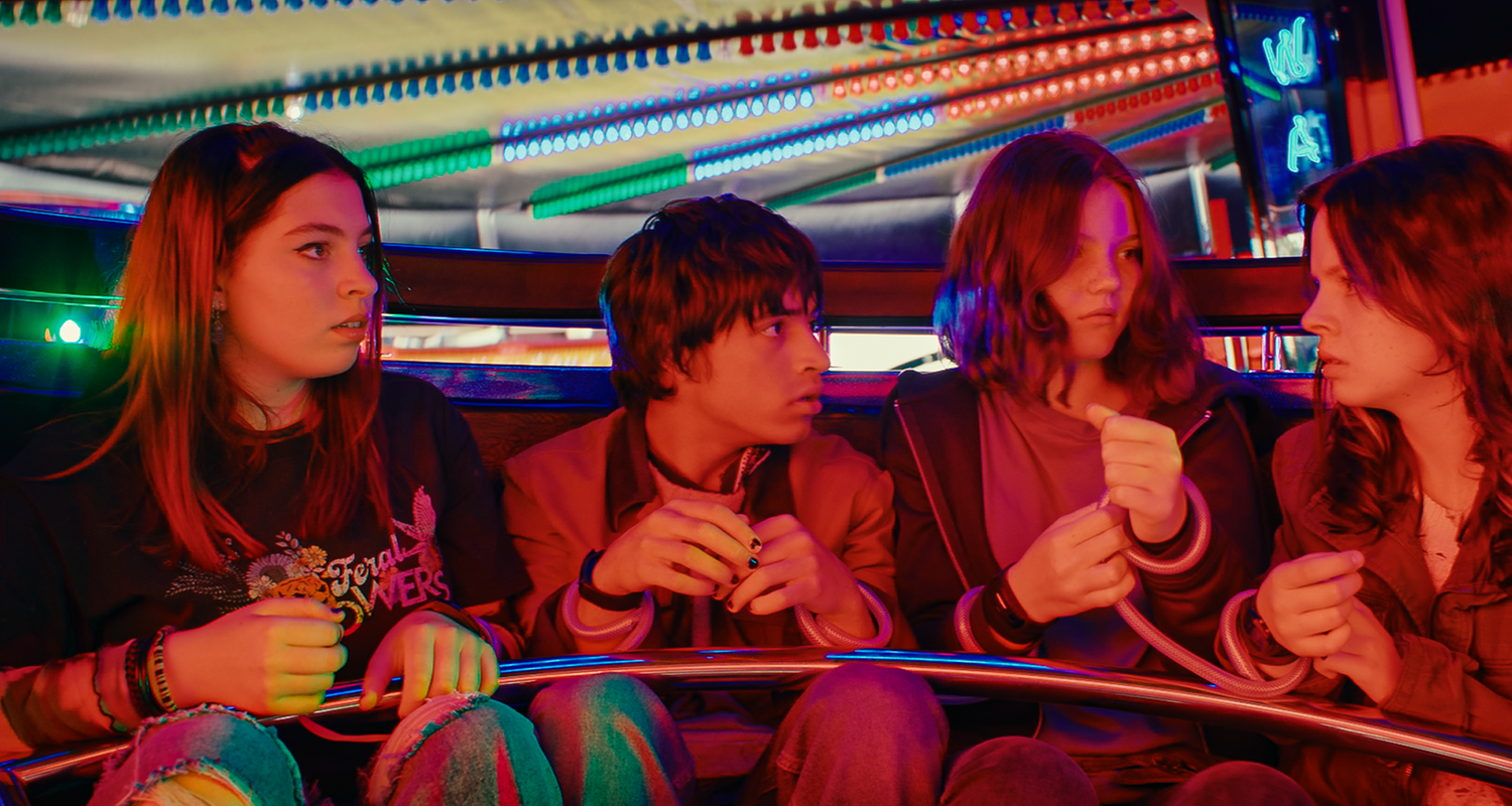 Auf einem Jahrmarkt in einem Wagen eines Karussells sitzen Kaz, Jay, Isabel und Emma eng nebeneinander. Sie sind an den Handgelenken mit durchsichtigen Schläuchen jeweils mit dem Sitznachbarn verbunden.