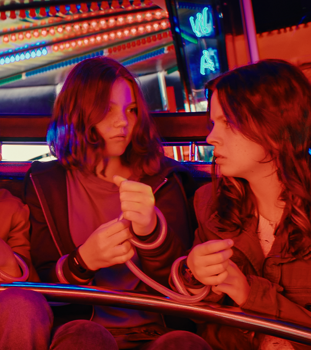 Auf einem Jahrmarkt in einem Wagen eines Karussells sitzen Kaz, Jay, Isabel und Emma eng nebeneinander. Sie sind an den Handgelenken mit durchsichtigen Schläuchen jeweils mit dem Sitznachbarn verbunden.