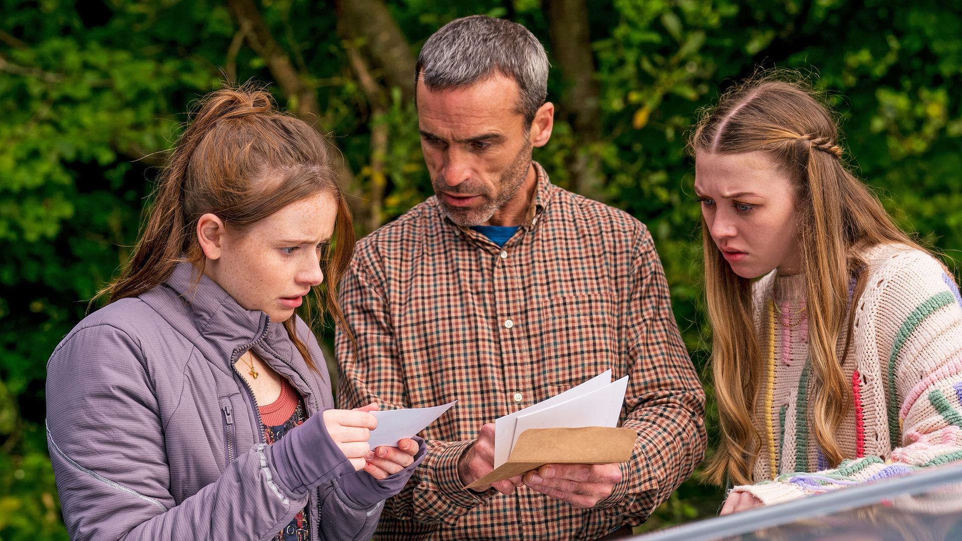 Draußen neben einem Auto stehen Maeves Vater Aiden, gespielt von Dara Devaney, und Maeve, gespielt von Skye McClenaghan. Auch Bea, dargestellt von Aoife Hughes, steht neben ihnen. Aiden und Bea halten Fotos in den Händen. Aiden schaut zu Bea, die konzentriert auf die Fotos blickt. Maeve sieht von der Seite aus mit angestrengtem Gesicht zu.