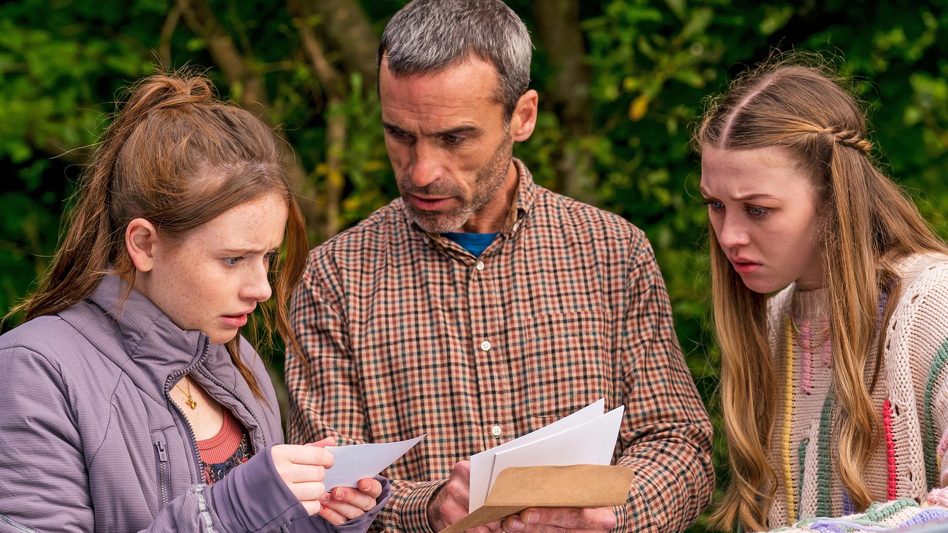 Draußen neben einem Auto stehen Maeves Vater Aiden, gespielt von Dara Devaney, und Maeve, gespielt von Skye McClenaghan. Auch Bea, dargestellt von Aoife Hughes, steht neben ihnen. Aiden und Bea halten Fotos in den Händen. Aiden schaut zu Bea, die konzentriert auf die Fotos blickt. Maeve sieht von der Seite aus mit angestrengtem Gesicht zu.