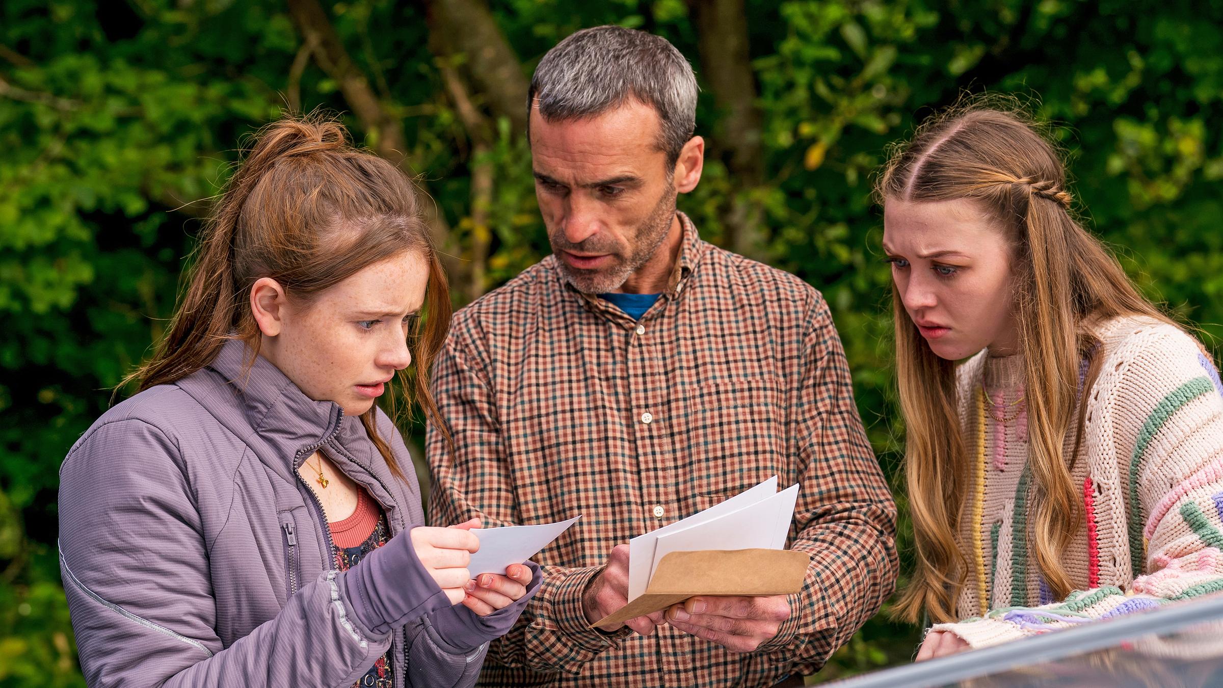 Draußen neben einem Auto stehen Maeve Vater Aiden, gespielt von Dara Devaney, und Maeve, gespielt von Skye McClenaghan. Auch Bea, dargestellt von Aoife Hughes, steht neben ihnen. Aiden und Bea halten Fotos in den Händen. Aiden schaut zu Bea, die konzentriert auf die Fotos blickt. Maeve sieht von der Seite aus mit angestrengtem Gesicht zu.