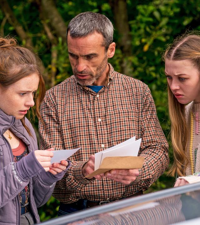 Draußen neben einem Auto stehen Maeves Vater Aiden, gespielt von Dara Devaney, und Maeve, gespielt von Skye McClenaghan. Auch Bea, dargestellt von Aoife Hughes, steht neben ihnen. Aiden und Bea halten Fotos in den Händen. Aiden schaut zu Bea, die konzentriert auf die Fotos blickt. Maeve sieht von der Seite aus mit angestrengtem Gesicht zu.