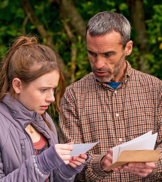 Draußen neben einem Auto stehen Maeves Vater Aiden, gespielt von Dara Devaney, und Maeve, gespielt von Skye McClenaghan. Auch Bea, dargestellt von Aoife Hughes, steht neben ihnen. Aiden und Bea halten Fotos in den Händen. Aiden schaut zu Bea, die konzentriert auf die Fotos blickt. Maeve sieht von der Seite aus mit angestrengtem Gesicht zu.