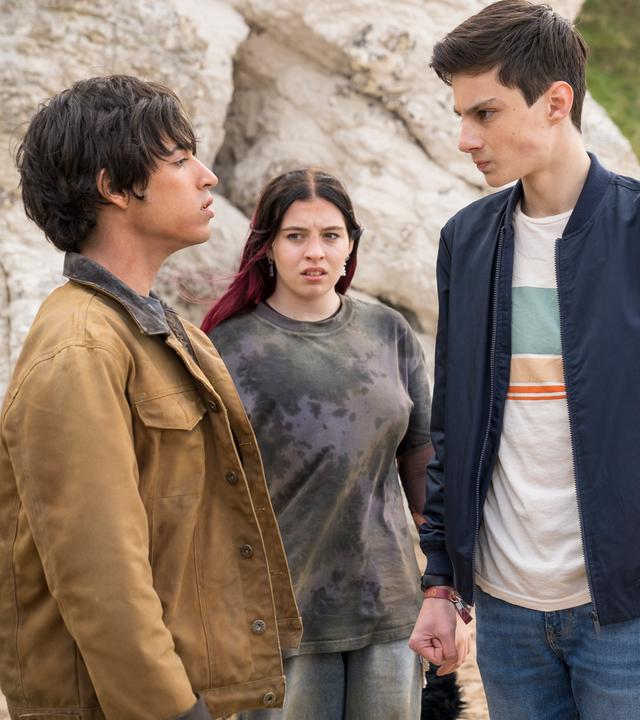 Am Strand vor hellem Felsen stehen sich Louis, gespielt von Oliver Cunliffe, und Jay, gespielt von Noah Manzoor, gegenüber. Louis ist voller Wut mit angespanntem Gesicht und geballten Fäusten. Jay schaut ihn herausfordernd an. Die beiden werden von Kaz, gespielt von Maiya Silveston, und Isabel, gespielt vo Alis Waters, beobachtet. Kaz schaut mit einem verächtlichen Blick zu Jay.