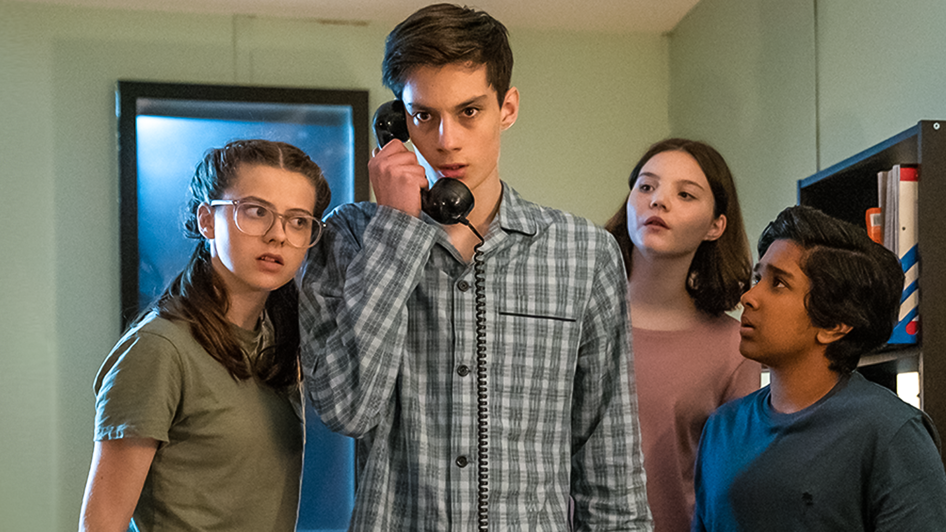 In einem Büroraum bei Nacht stehen Monika, gespielt von Lucy Chambers, Isabel, gespielt von Alis Waters, und Glen, dargestellt von Krish Misra, um Louis, Darsteller Oliver Cunliffe, herum. Louis hält einen alten schwarzen Telefonhörer in der Hand. Die drei schauen neugierig und aufmerksam zu Louis, während dieser ernst nach vorne schaut. Alle tragen Schlafanzüge.