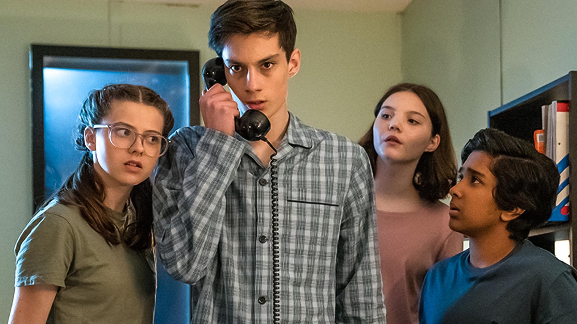 In einem Büroraum bei Nacht stehen Monika, gespielt von Lucy Chambers, Isabel, gespielt von Alis Waters, und Glen, dargestellt von Krish Misra, um Louis, Darsteller Oliver Cunliffe, herum. Louis hält einen alten schwarzen Telefonhörer in der Hand. Die drei schauen neugierig und aufmerksam zu Louis, während dieser ernst nach vorne schaut. Alle tragen Schlafanzüge.