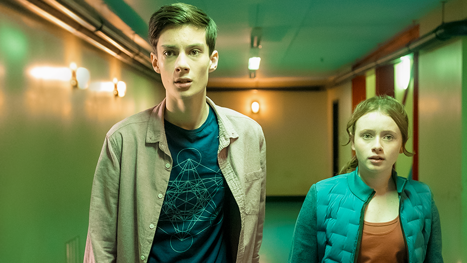 In einem grünlich beleuchteten Flur stehen Bea, gespielt von Aoife Hughes, und Louis, gespielt von Oliver Cunliffe, nebeneinander. Beide schauen ängstlich und angespannt nach vorne.