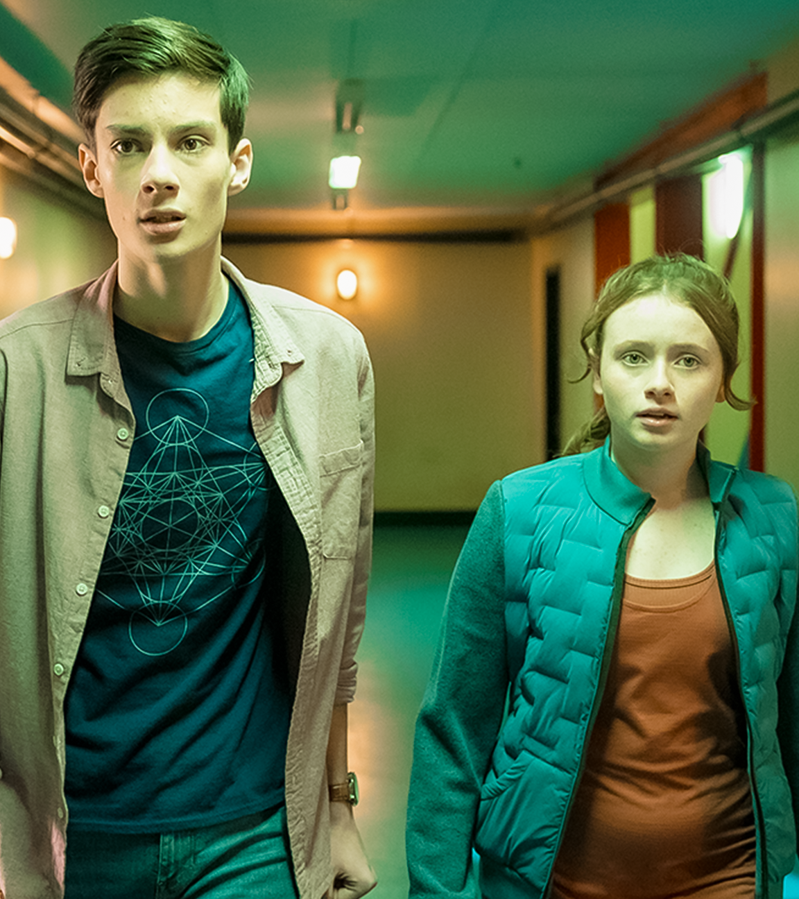 In einem grünlich beleuchteten Flur stehen Bea, gespielt von Aoife Hughes, und Louis, gespielt von Oliver Cunliffe, nebeneinander. Beide schauen ängstlich und angespannt nach vorne.