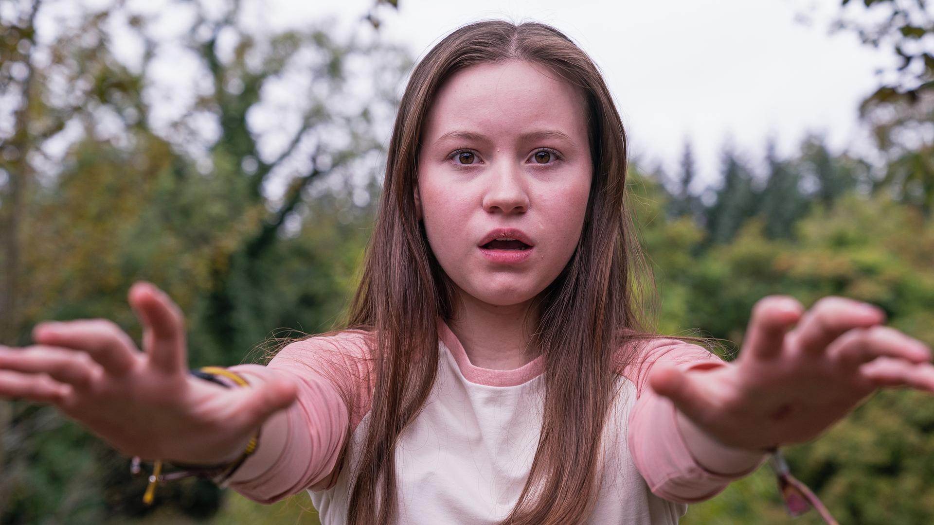 Meg, gespielt von Katy Byrne, steht draußen vor Bäumen und Büschen. Der Wind weht ihr ins Gesicht und sie schaut angestrengt nach vorne. Ihr Blick sieht besorgt aus.