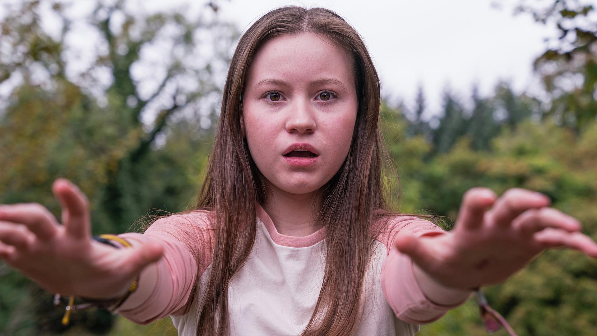 Meg, gespielt von Katy Byrne, steht draußen vor Bäumen und Büschen. Der Wind weht ihr ins Gesicht und sie schaut angestrengt nach vorne. Ihr Blick sieht besorgt aus.