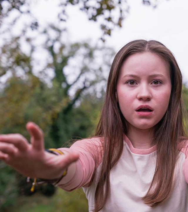Meg, gespielt von Katy Byrne, steht draußen vor Bäumen und Büschen. Der Wind weht ihr ins Gesicht und sie schaut angestrengt nach vorne. Ihr Blick sieht besorgt aus.