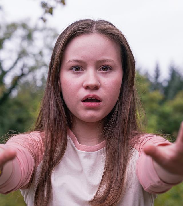 Meg, gespielt von Katy Byrne, steht draußen vor Bäumen und Büschen. Der Wind weht ihr ins Gesicht und sie schaut angestrengt nach vorne. Ihr Blick sieht besorgt aus.