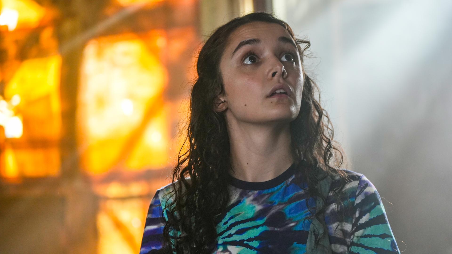 Addie steht in einem abgedunkelten Buchladen. Sie befindet sich im Zentrum des Bildes, ist frontal zu sehen und macht einen erfrüchtigen, beeindruckten Gesichtsausdruck.