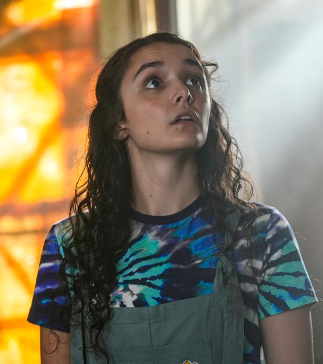 Addie steht in einem abgedunkelten Buchladen. Sie befindet sich im Zentrum des Bildes, ist frontal zu sehen und macht einen erfrüchtigen, beeindruckten Gesichtsausdruck.