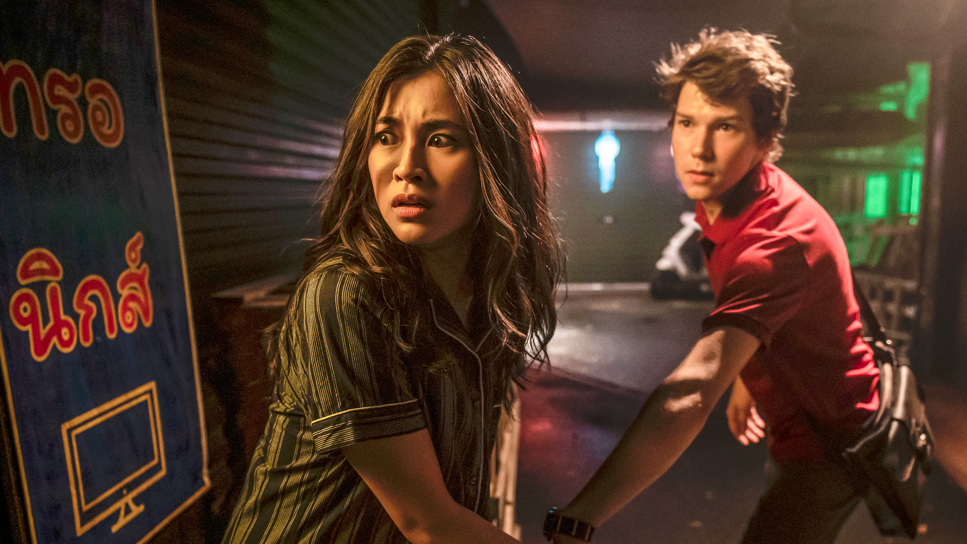 Kyra und Darra stehen mit erschrockenem Blick in einer dunklen, schmalen Straße Bangkogs bei Nacht. Darra zieht Kyra mit sich, als wären sie auf der Flucht. Beide scheinen auf mögliche Verfolger zu blicken.