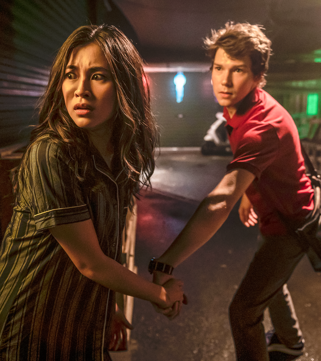 Kyra und Darra stehen mit erschrockenem Blick in einer dunklen, schmalen Straße Bangkogs bei Nacht. Darra zieht Kyra mit sich, als wären sie auf der Flucht. Beide scheinen auf mögliche Verfolger zu blicken.