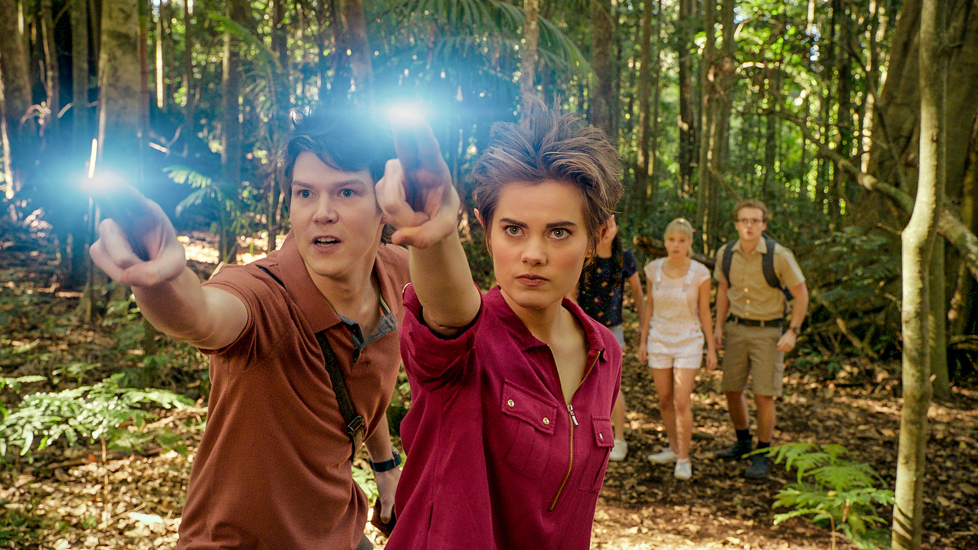 Darra und Imogen stehen nebeneinander in einem dichten Wald auf einer Lichtung und strecken beide zwei leuchtende Finger ihrer rechten Hand nach vorne. Ihre Freunde Kyra, Lily und Peter stehen in einiger Entfernung hinter ihnen. Alle schauen gebannt nach vorne.