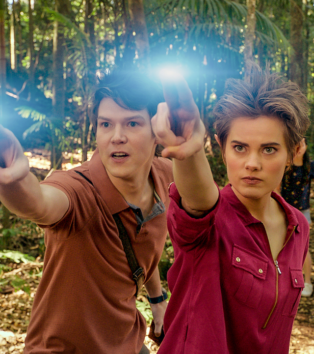 Darra und Imogen stehen nebeneinander in einem dichten Wald auf einer Lichtung und strecken beide zwei leuchtende Finger ihrer rechten Hand nach vorne. Ihre Freunde Kyra, Lily und Peter stehen in einiger Entfernung hinter ihnen. Alle schauen gebannt nach vorne.