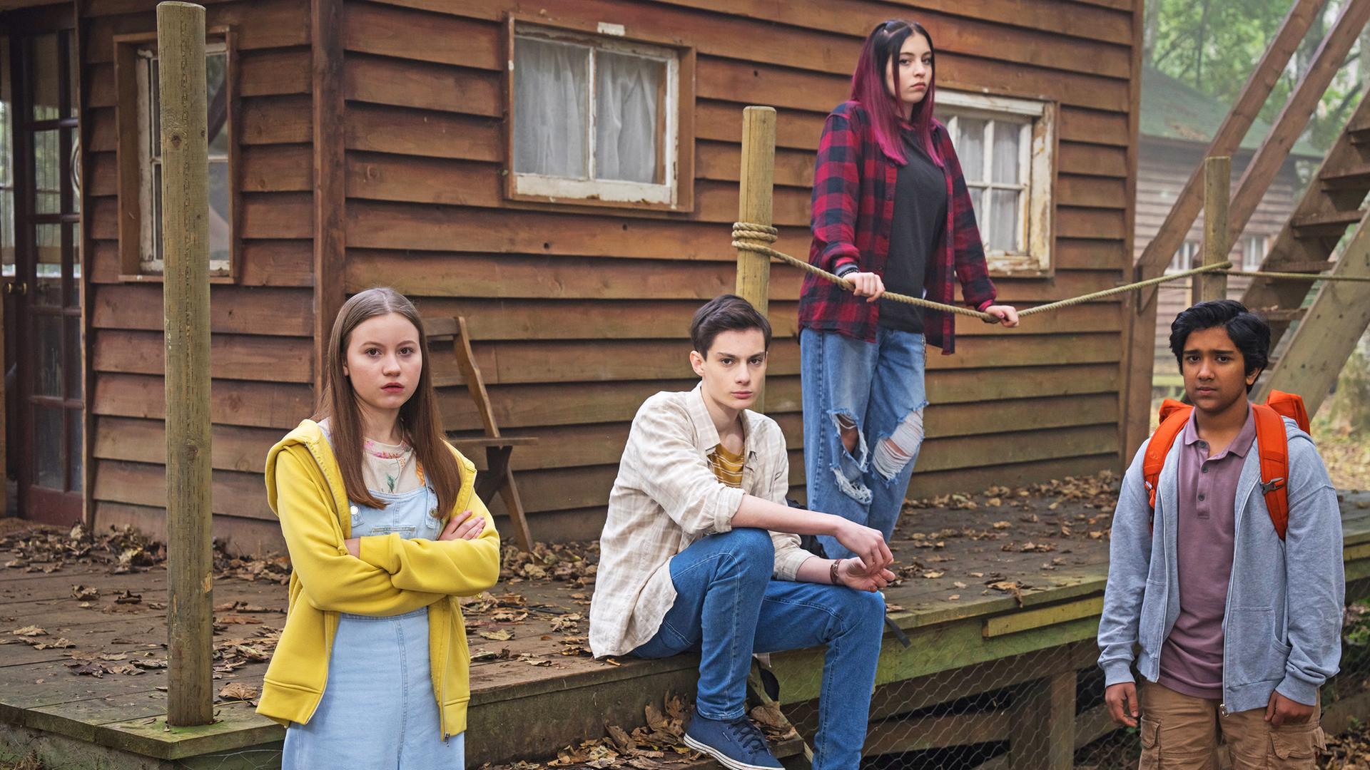 Wieder im Wald im Camp-Silverpoint: Die Libellen-Gruppe steht vor einer der Holzhütten und schaut sich verwirrt und skeptisch um.