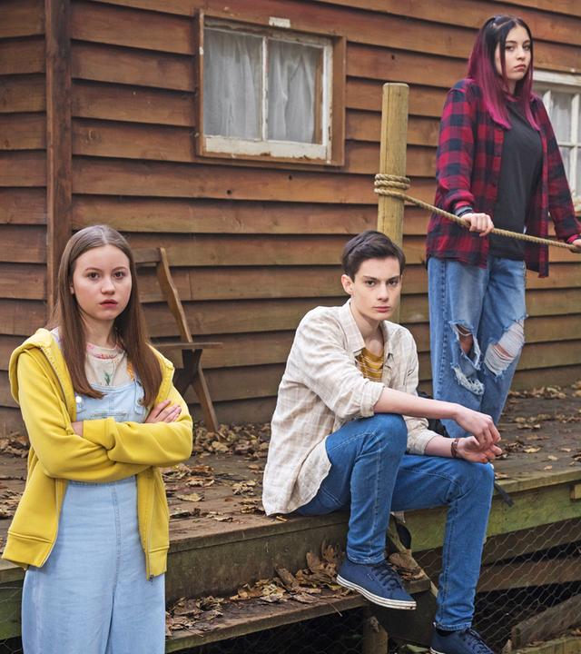 Wieder im Wald im Camp-Silverpoint: Die Libellen-Gruppe steht vor einer der Holzhütten und schaut sich verwirrt und skeptisch um.