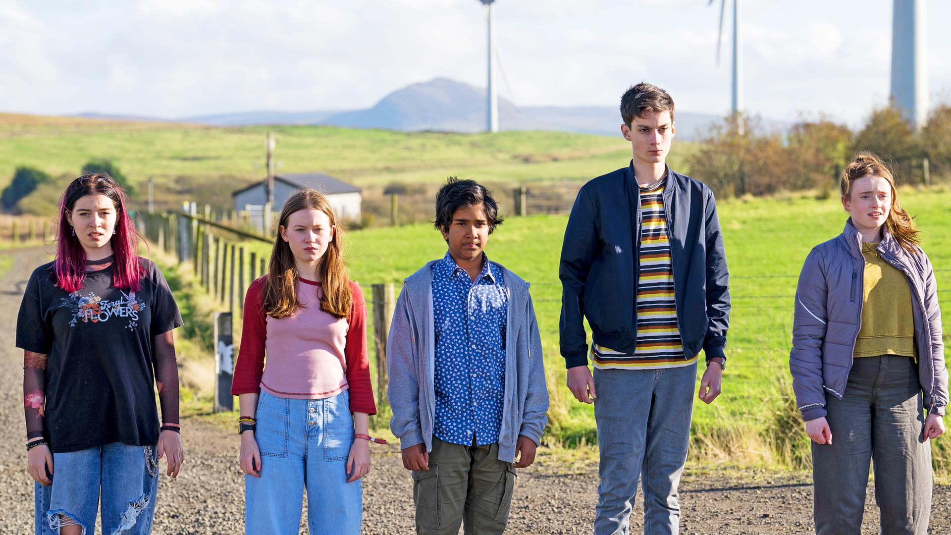 Kaz, Meg, Glen und Louis stehen nebeneinander auf einem Feldweg, mit Feldern und mehreren Windrädern im Hintergrund. Die Sonne scheint, sie schauen alle etwas verunsichert nach vorne. Glen hat ein leichtes Lächeln um den Mund herum.