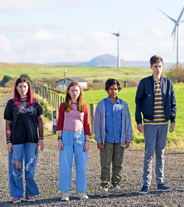 Kaz, Meg, Glen und Louis stehen nebeneinander auf einem Feldweg, mit Feldern und mehreren Windrädern im Hintergrund. Die Sonne scheint, sie schauen alle etwas verunsichert nach vorne. Glen hat ein leichtes Lächeln um den Mund herum.