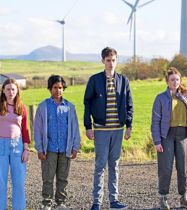 Kaz, Meg, Glen und Louis stehen nebeneinander auf einem Feldweg, mit Feldern und mehreren Windrädern im Hintergrund. Die Sonne scheint, sie schauen alle etwas verunsichert nach vorne. Glen hat ein leichtes Lächeln um den Mund herum.