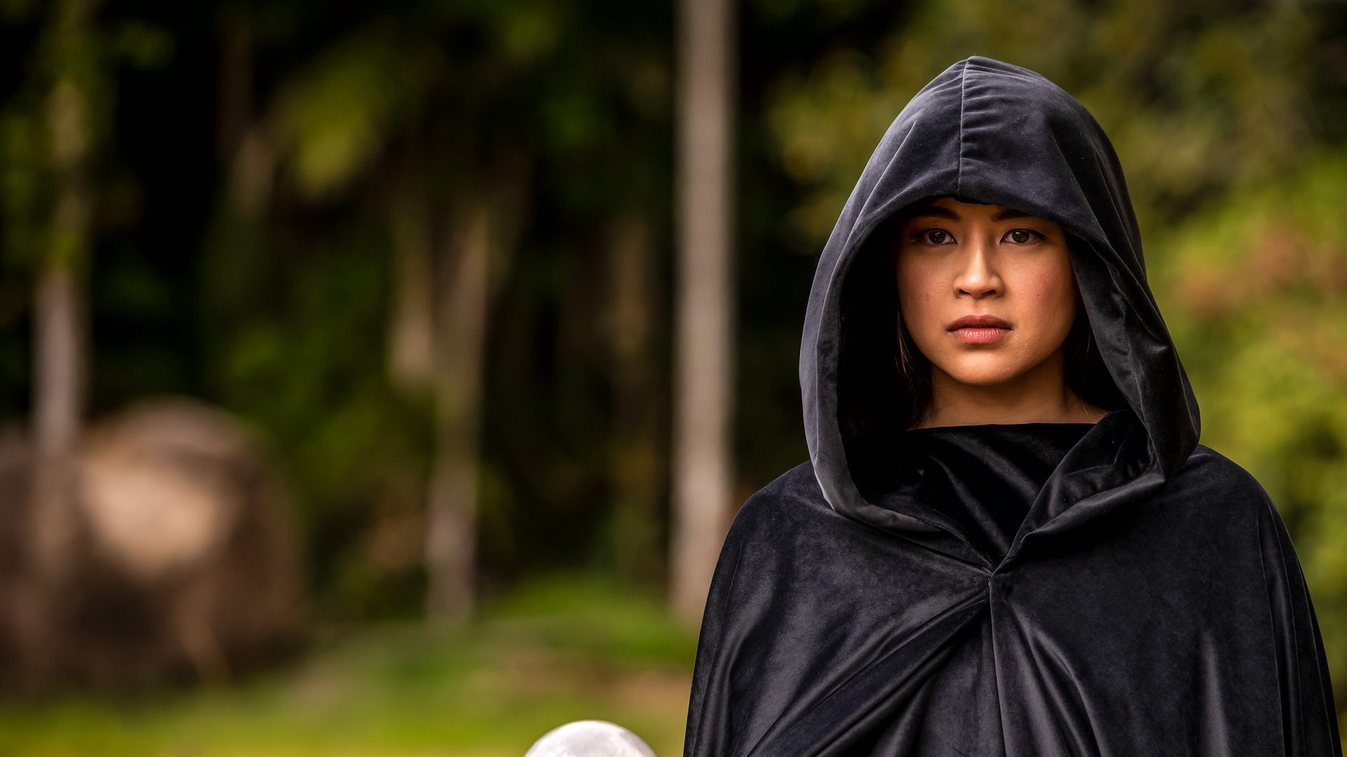 Kyra steht, in einen schwarzen Kapuzenumhang gehüllt, in einem Wald. Sie hält in der rechten Hand eine weiße Kugel und blickt ernst nach vorne.