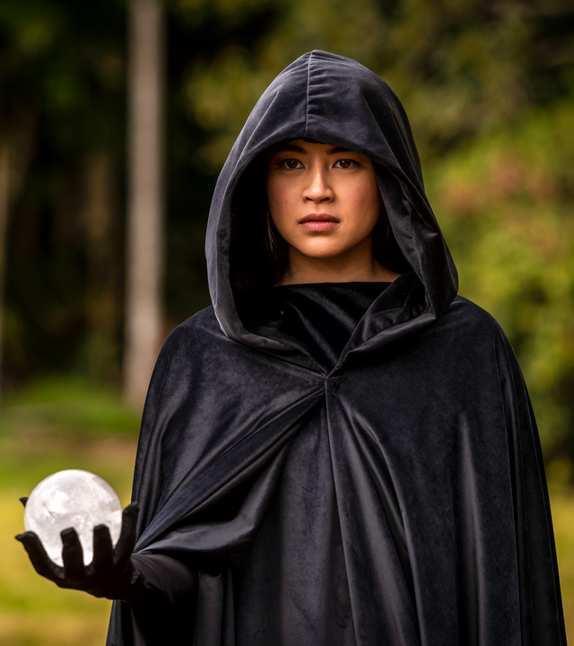 Kyra steht, in einen schwarzen Kapuzenumhang gehüllt, in einem Wald. Sie hält in der rechten Hand eine weiße Kugel und blickt ernst nach vorne.