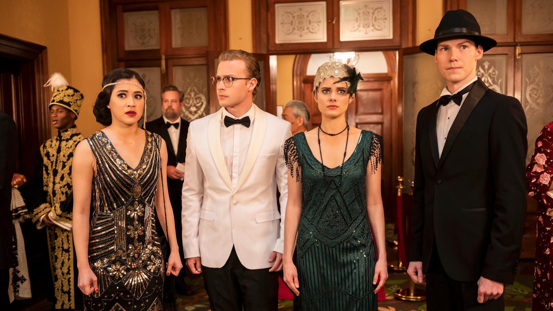 Kyra, Peter, Imogen und Darra stehen in festlicher Kleidung in der Mode von 1919 nebeneinander in einer Empfangshalle zwischen anderen Gästen. Sie schauen sich angespannt um.