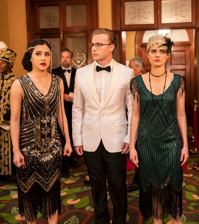 Kyra, Peter, Imogen und Darra stehen in festlicher Kleidung in der Mode von 1919 nebeneinander in einer Empfangshalle zwischen anderen Gästen. Sie schauen sich angespannt um.