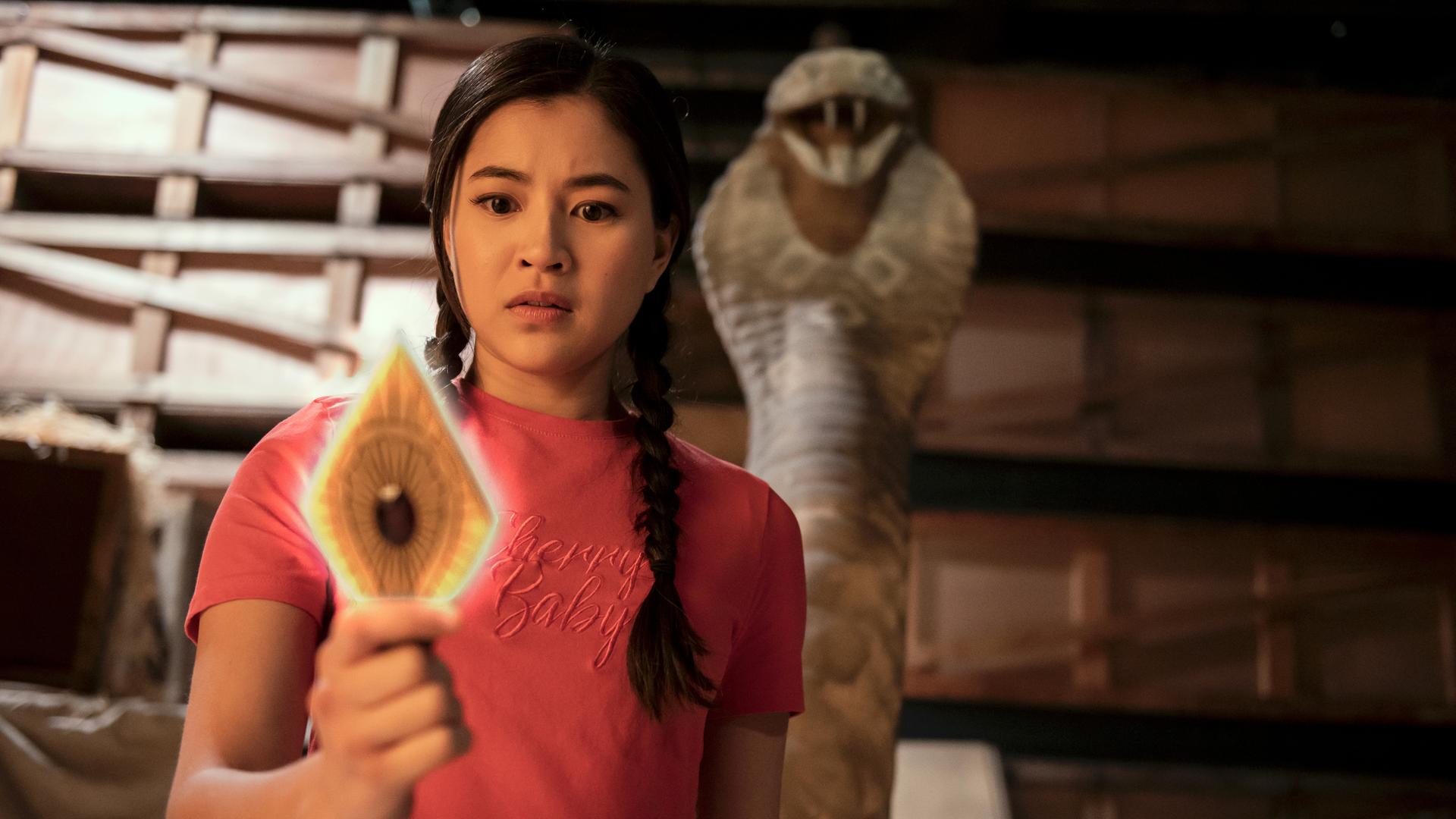 Kyra steht in einem dunklen Lagerraum vor Holzregalen. Hinter ihr ragt eine steinerne Schlange auf. Kyra hält eine leuchtende goldfarbene Metallplatte in Rautenform und mit einem roten Edelstein in der Mitte in der rechten Hand. Sie schaut verwundert auf die Platte.