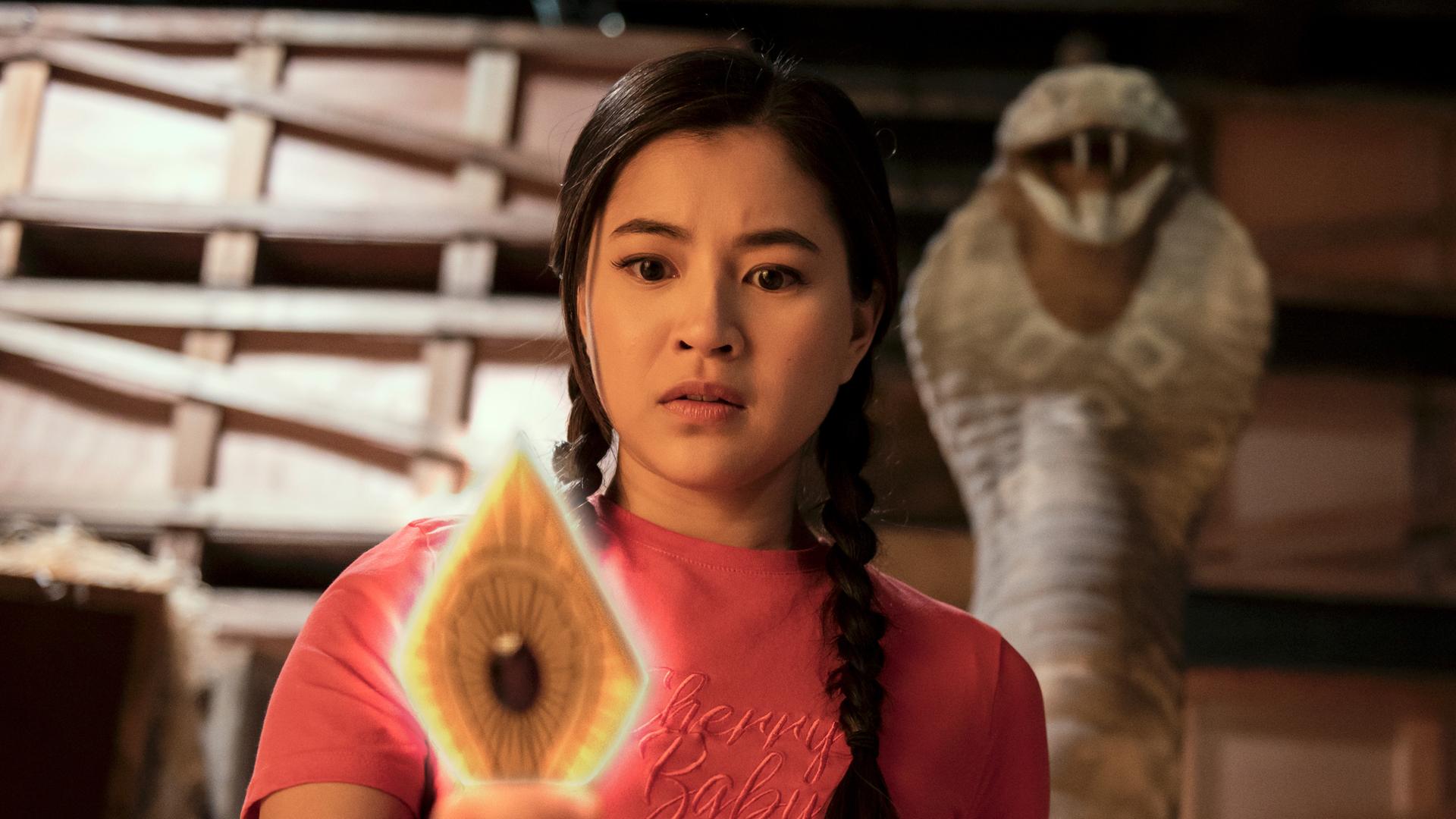 Kyra steht in einem dunklen Lagerraum vor Holzregalen. Hinter ihr ragt eine steinerne Schlange auf. Kyra hält eine leuchtende goldfarbene Metallplatte in Rautenform und mit einem roten Edelstein in der Mitte in der rechten Hand. Sie schaut verwundert auf die Platte.