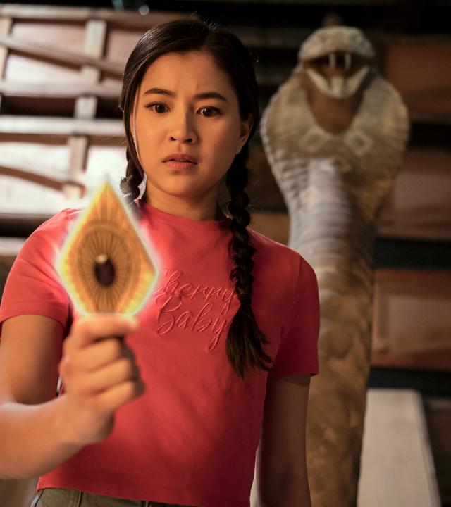 Kyra steht in einem dunklen Lagerraum vor Holzregalen. Hinter ihr ragt eine steinerne Schlange auf. Kyra hält eine leuchtende goldfarbene Metallplatte in Rautenform und mit einem roten Edelstein in der Mitte in der rechten Hand. Sie schaut verwundert auf die Platte.