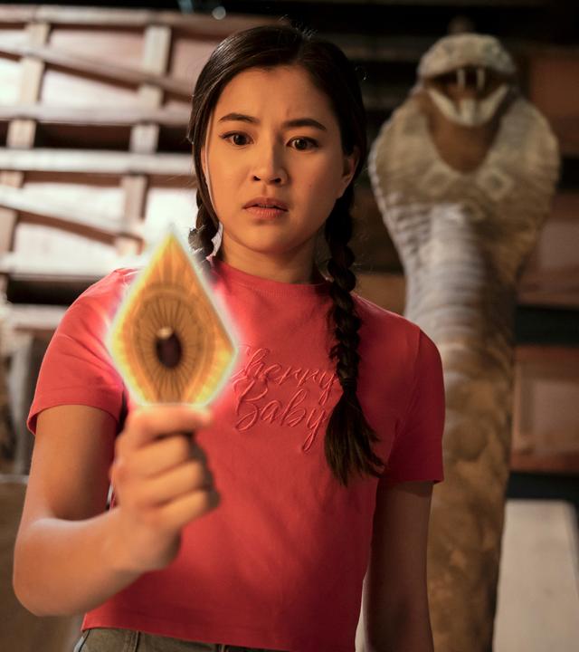 Kyra steht in einem dunklen Lagerraum vor Holzregalen. Hinter ihr ragt eine steinerne Schlange auf. Kyra hält eine leuchtende goldfarbene Metallplatte in Rautenform und mit einem roten Edelstein in der Mitte in der rechten Hand. Sie schaut verwundert auf die Platte.