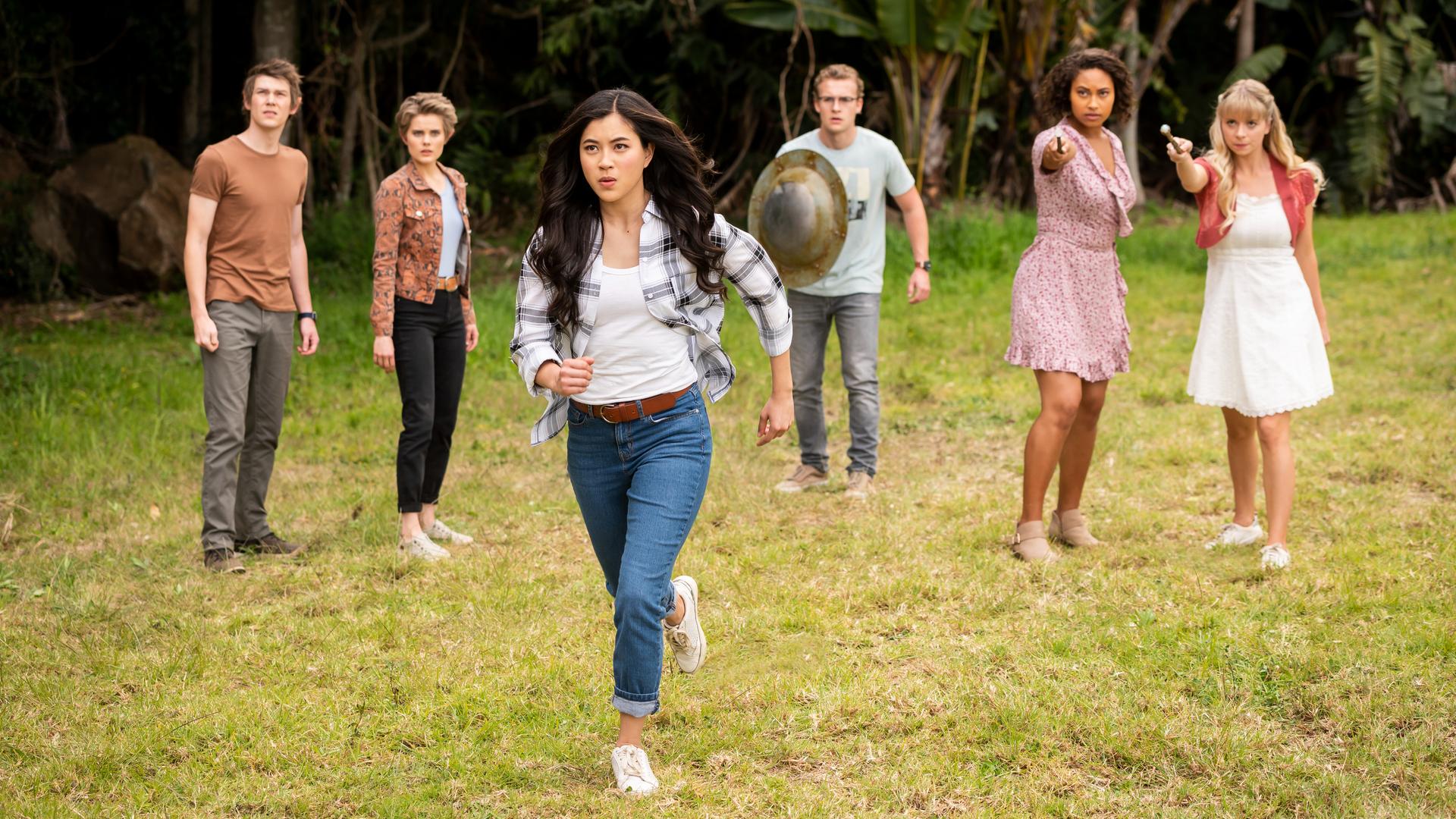 Darra, Imogen, Peter, Ruksy und Lily stehen hinter Kyra auf einer Wiese, die von dichtem Wald umgeben ist. Kyra beginnt, auf ein vor ihr liegendes Ziel loszurennen. Ruksy und Lily strecken ihre Zauberstäbe nach vorne, Peter hält ein Metallschild in der rechten Hand vor dem Körper. Alle sind angespannt und schauen ernst nach vorne.