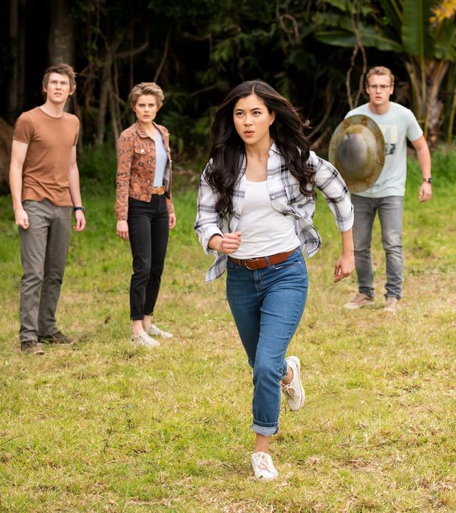 Darra, Imogen, Peter, Ruksy und Lily stehen hinter Kyra auf einer Wiese, die von dichtem Wald umgeben ist. Kyra beginnt, auf ein vor ihr liegendes Ziel loszurennen. Ruksy und Lily strecken ihre Zauberstäbe nach vorne, Peter hält ein Metallschild in der rechten Hand vor dem Körper. Alle sind angespannt und schauen ernst nach vorne.