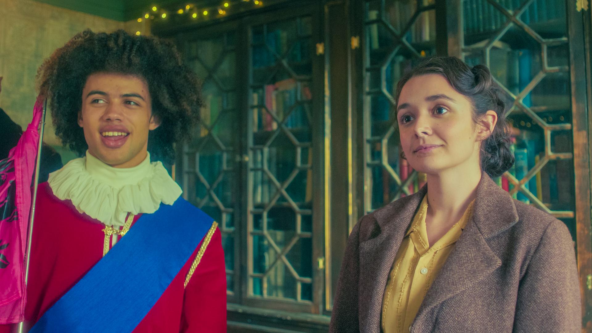 Im Buchladen stehen Frank und Addie nebeneinander vor einem verglasten Bücherschrank. Frank, der als historischer Stadtführer verkleidet ist, lächelt mit offenem Mund, und Addie schaut mit stolzem Blick und leicht erhobenem Kopf nach vorne.