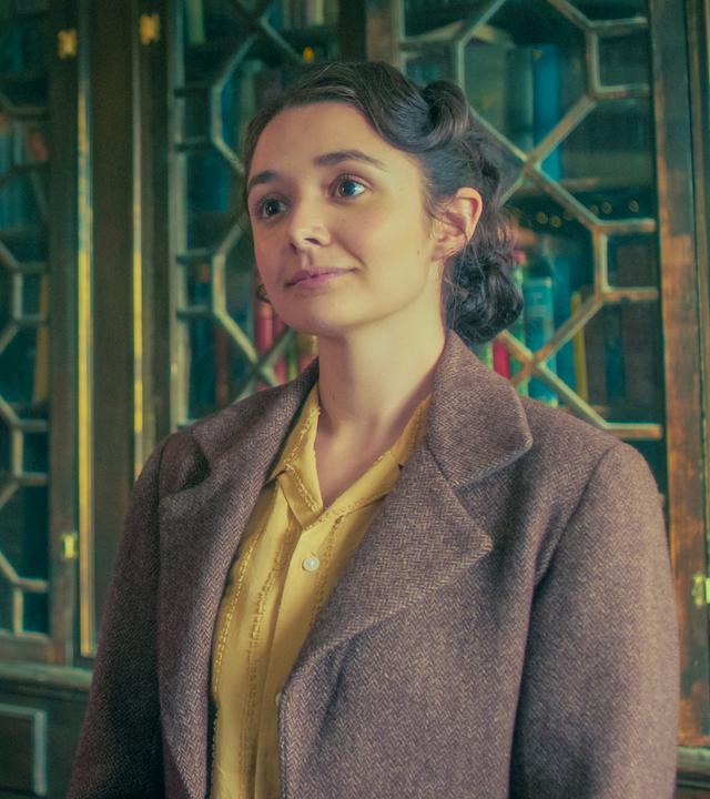 Im Buchladen stehen Frank und Addie nebeneinander vor einem verglasten Bücherschrank. Frank, der als historischer Stadtführer verkleidet ist, lächelt mit offenem Mund, und Addie schaut mit stolzem Blick und leicht erhobenem Kopf nach vorne.