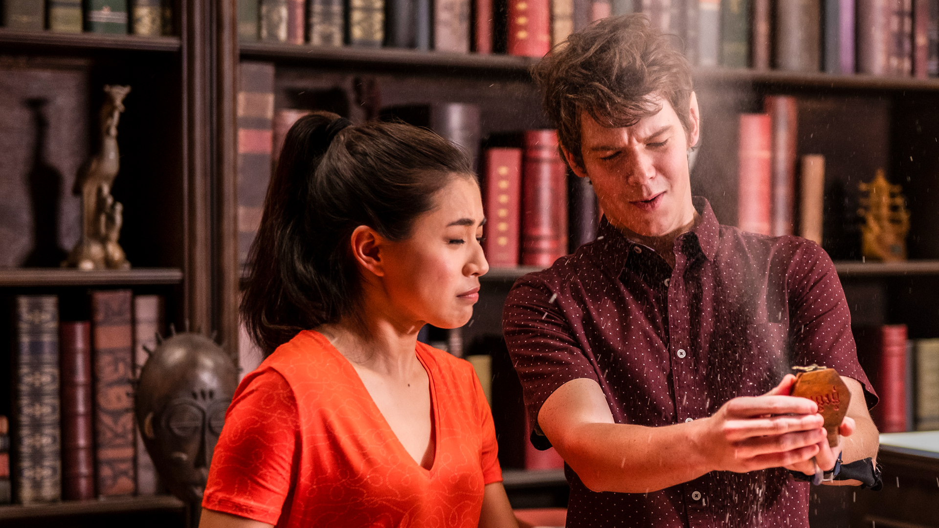 Kyra und Darra stehen vor einer Bücherwand in der Bibliothek. Vor ihnen stehen geöffnete Kisten. Darra öffnet gerade eine kleine Dose, aus der Staub aufwirbelt. Kyra und er schließen die Augen und drehen sich leicht weg von der Dose.