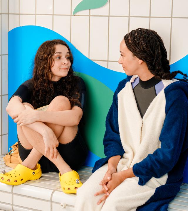 Addie und Audrey sitzen nebeneinander auf einer Bank im Schwimmbad, angelehnt an eine Wand mit blau-grünem Wellenmuster. Addie hat ihre Beine angezogen und ihre Arme darum gelegt. Sie trägt Badekleidung und gelbe Badeschuhe. Sie schaut etwas verschüchtert zu ihrer besten Freundin. Audrey trägt ein Haikostüm. Ihre Hände liegen ineinander verschränkt in ihrem Schoß. Sie schaut lächelnd zu Addie.