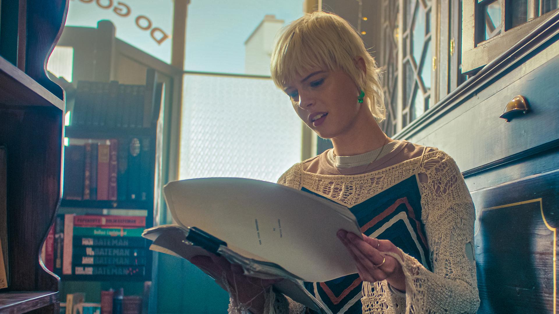 Im Buchladen sitzt Bonnie zwischen zwei Bücherschränken im Schneidersitz auf dem Boden. Sie hält ein geöffnetes Manuskript in ihren Händen und schaut mit erstaunt-interessiertem Blick hinein. Direkt vor ihr steht eine offene Kiste. Hinter ihr steht ein Regal mit Büchern.