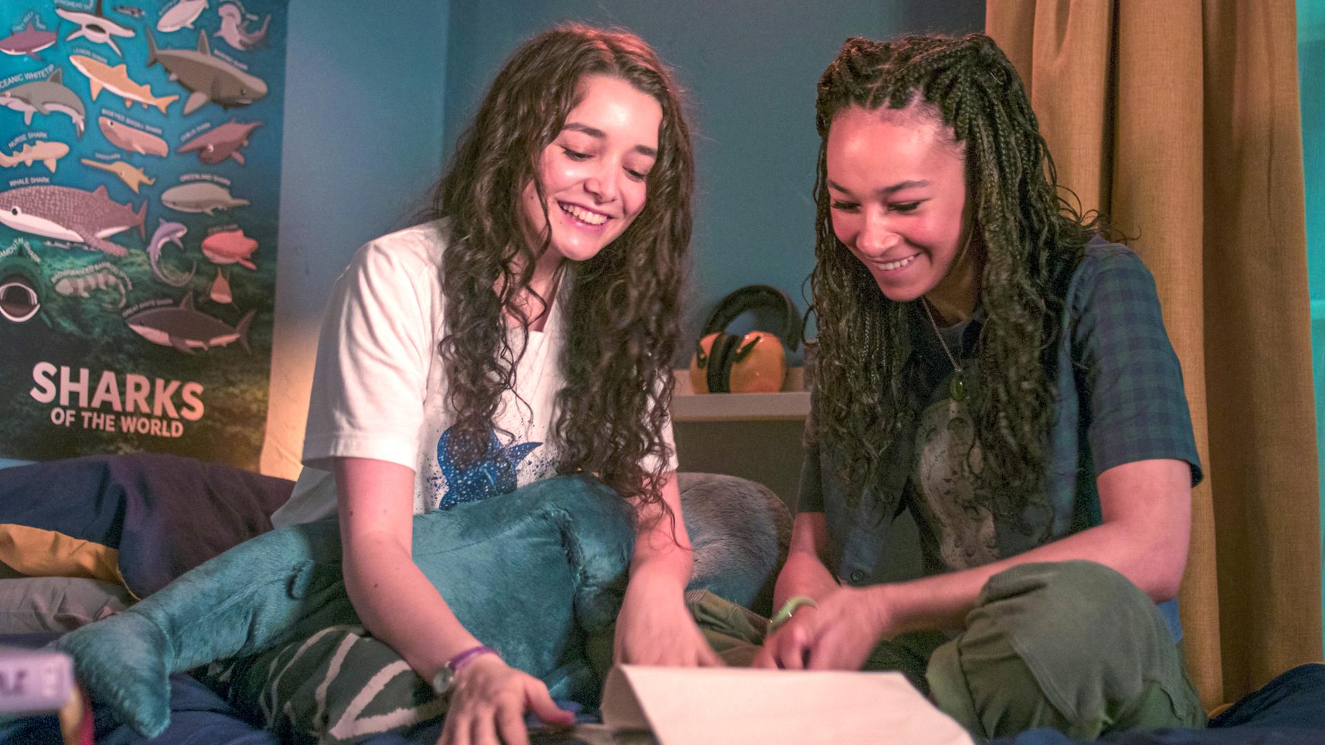In Addies abgedunkeltem Zimmer sitzen Addie und Audrey im Schneidersitz nebeneinander auf dem Bett. Sie haben ein geöffnetes Manuskript vor sich liegen und schauen beide lachend hinein. Im Hintergrund hängt ein Poster mit vielen verschiedenen Hai-Arten.
