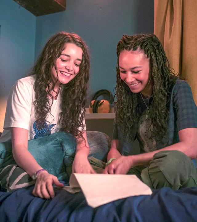 In Addies abgedunkeltem Zimmer sitzen Addie und Audrey im Schneidersitz nebeneinander auf dem Bett. Sie haben ein geöffnetes Manuskript vor sich liegen und schauen beide lachend hinein. Im Hintergrund hängt ein Poster mit vielen verschiedenen Hai-Arten.