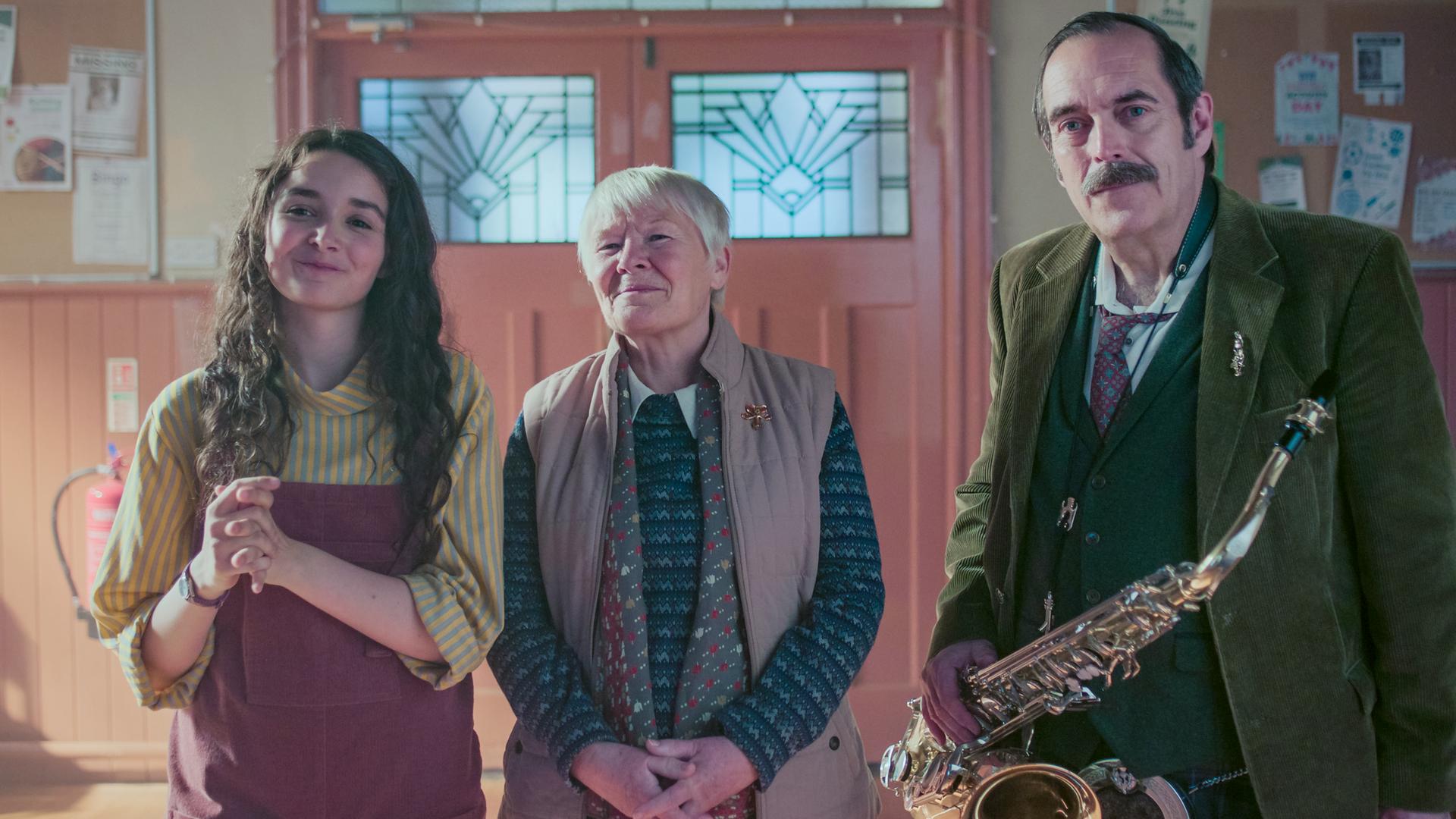 Addie, Miriam und Mr. Macintosh stehen nebeneinander und schauen begeistert lächelnd nach vorne. Mr. Macintosh hält ein Saxophon in seiner rechten Hand. Im Hintergrund ist eine geschlossene Flügeltür aus Holz, jeweils rechts und links neben der Tür hängen Pinnwände, an denen verschiedene Zettel befestigt sind.