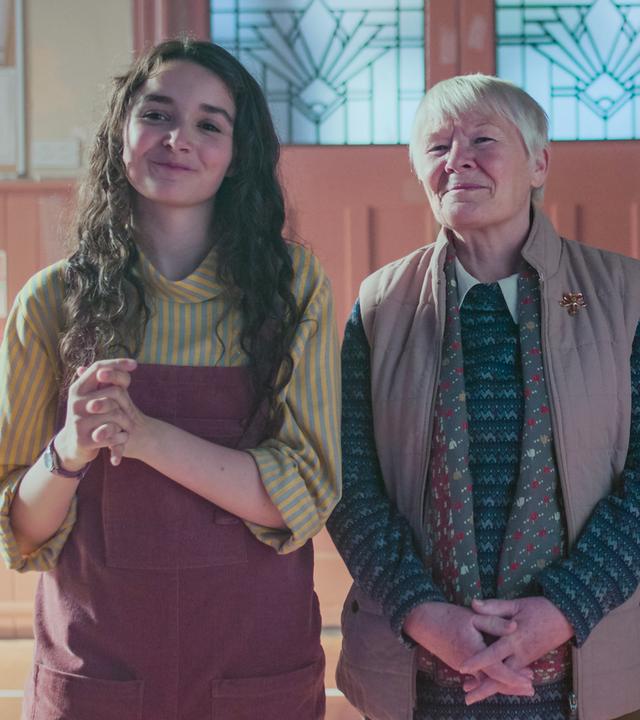 Addie, Miriam und Mr. Macintosh stehen nebeneinander und schauen begeistert lächelnd nach vorne. Mr. Macintosh hält ein Saxophon in seiner rechten Hand. Im Hintergrund ist eine geschlossene Flügeltür aus Holz, jeweils rechts und links neben der Tür hängen Pinnwände, an denen verschiedene Zettel befestigt sind.